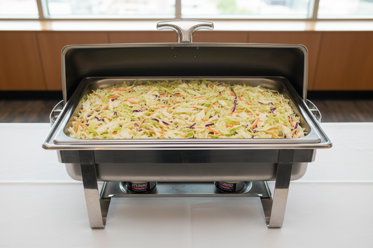 A high-quality corporate catering setup featuring a stainless steel chafing dish with the lid open, holding a full steam-table pan of fresh, creamy coleslaw. Finely shredded green cabbage, carrots, and purple cabbage evenly mixed and lightly coated in a smooth, glossy coleslaw dressing. The coleslaw is neatly leveled in the pan with clean edges, no spills. The chafing dish sits on a professional catering table with a white tablecloth, Sterno fuel visible below. Shot from a slightly elevated angle. Bright, c